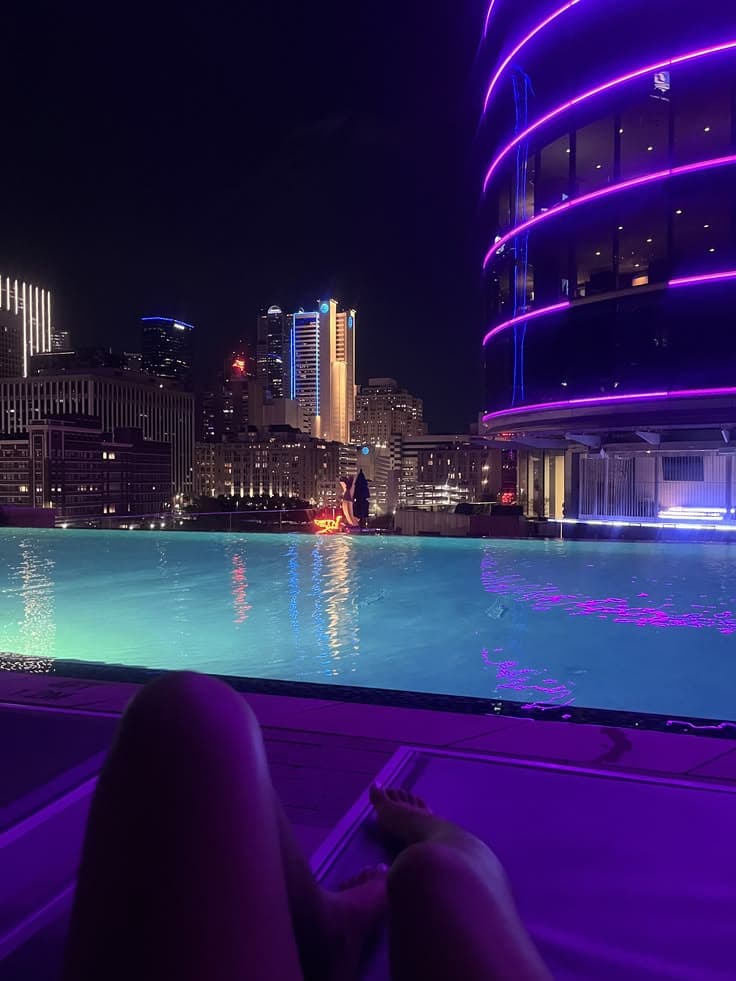 a person laying on the edge of a swimming pool at night with city lights in the background
