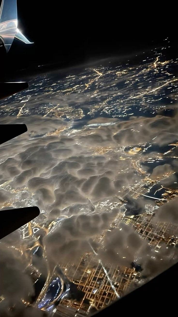 an airplane wing flying over the city lights at night in the sky with clouds below