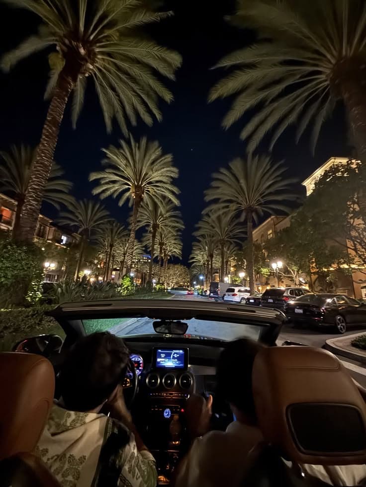 two people driving down the street at night with palm trees and buildings in the backgroun