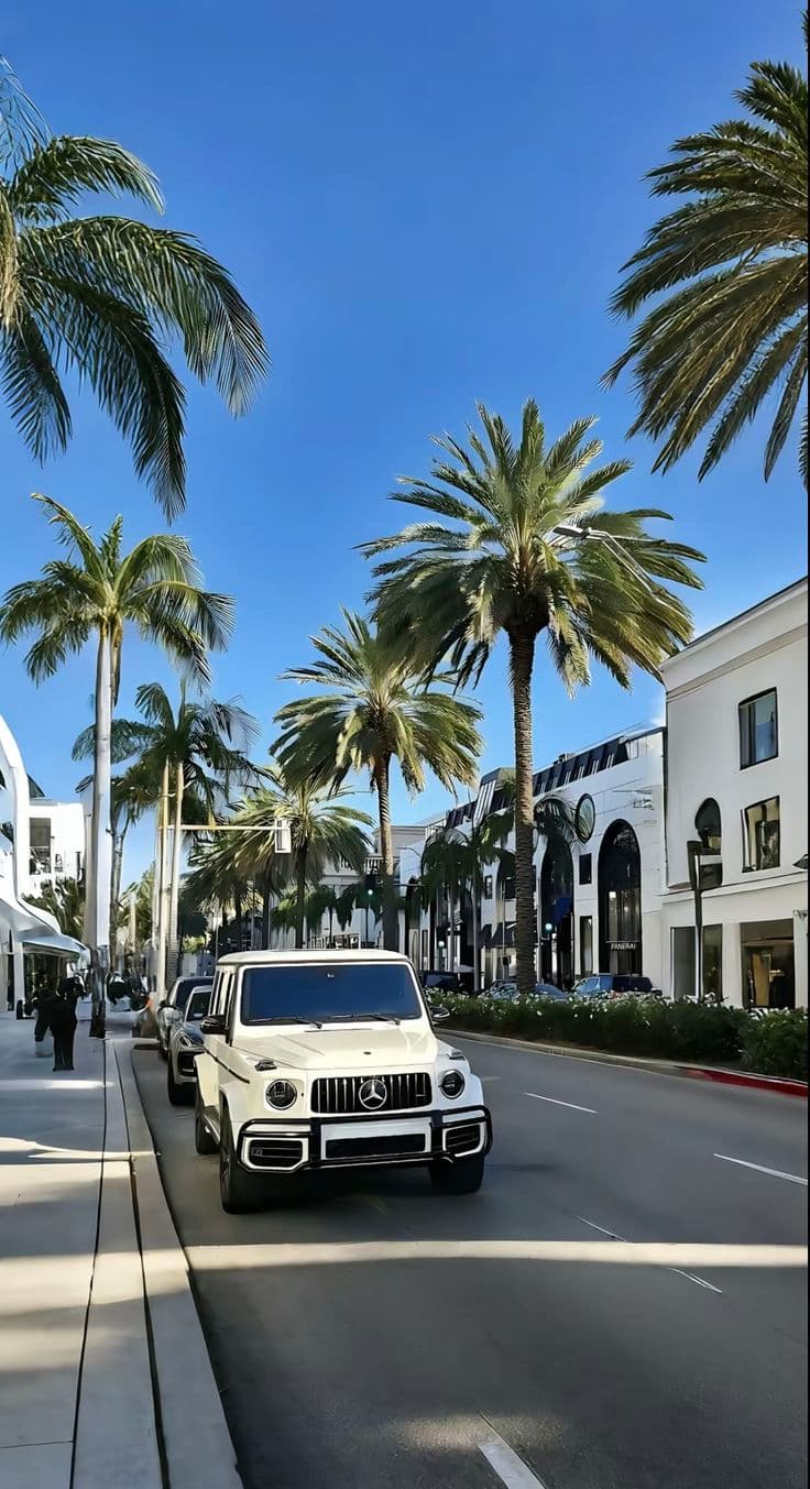 a white car driving down a street next to tall palm tree lined buildings on both sides