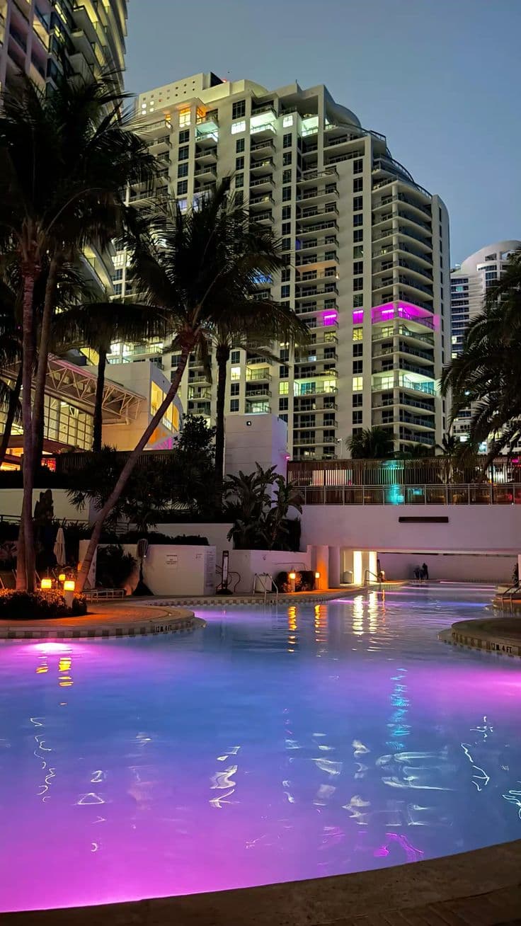 an outdoor swimming pool with palm trees and buildings in the background at night, lit up 