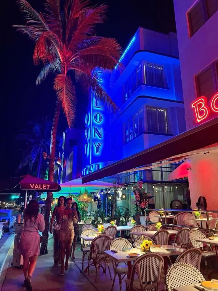 people are walking down the sidewalk in front of a building with neon lights and palm tree