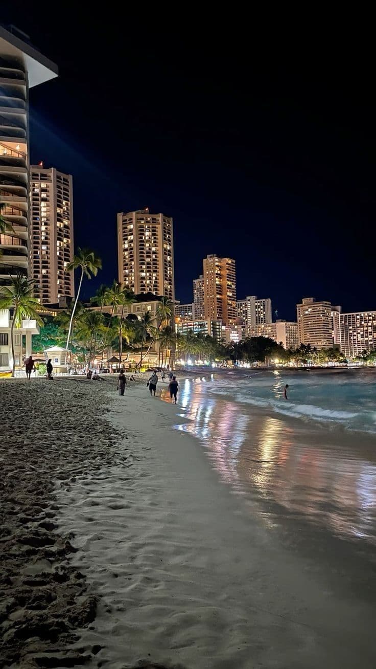 people walking on the beach at night with tall buildings in the backgrouds